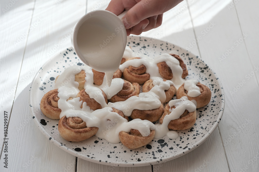 human hand pouring milk sauce or frosting on cinnamon swirls. selective focus. homemade cinnamon rolls with cream glaze. home cooking concept. human hand pouring milk sauce or frosting on cinnamon swirls. selective focus. homemade cinnamon rolls with cream glaze. home cooking concept.