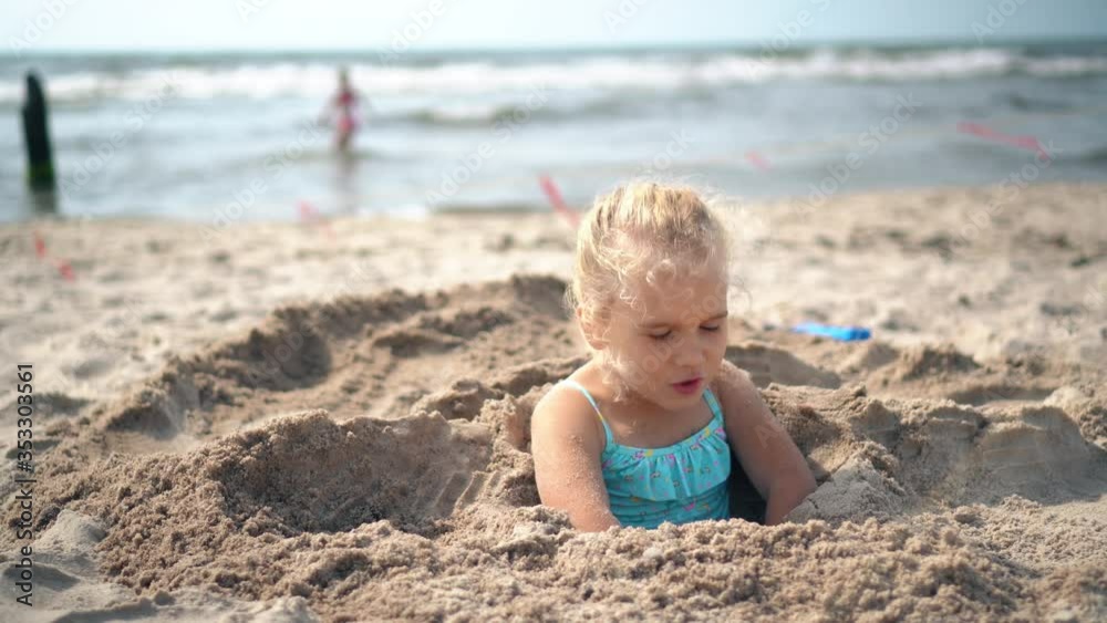 Blond adorable girl buried in sand try to break free. Blurred people and sea Blond adorable girl buried in sand try to break free. Blurred people and sea
