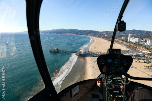 Helicopter cockpit flying on landscape and blue sky, with pilot arm driving in cabin. Spectacular view of ocean, curve road, mountains, Santa Monica Pier