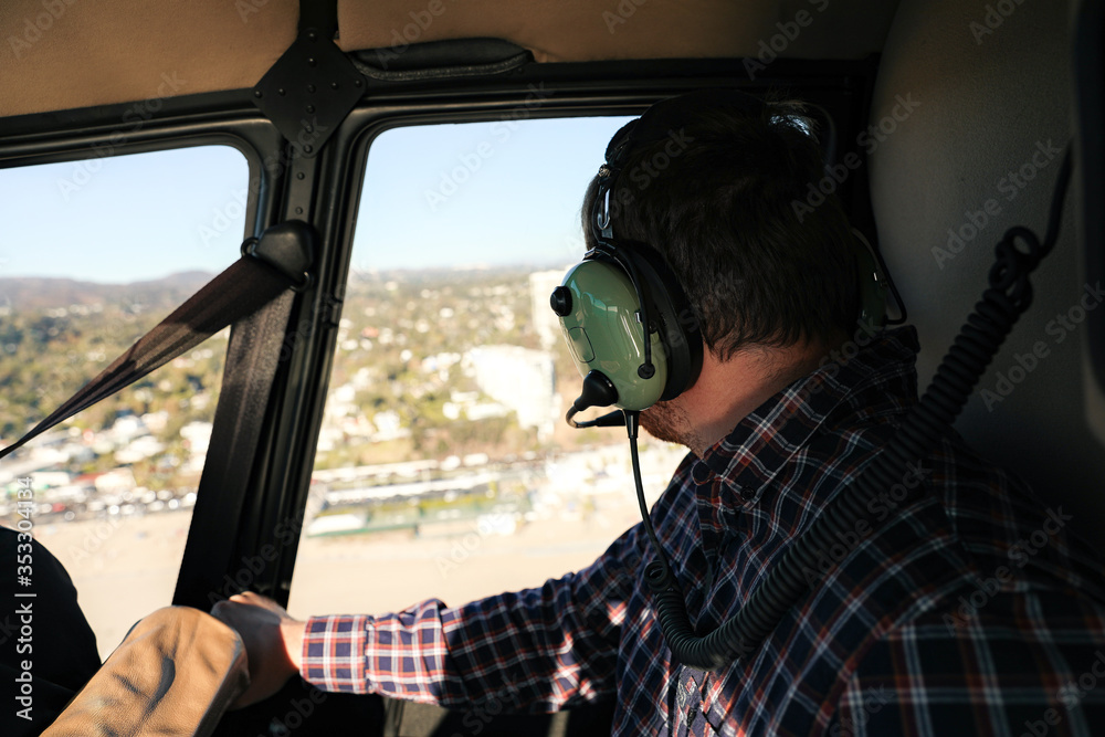 Serious confident young man in plaid shirt sitting in the helicopter cabin and wearing big headphones while checking the view through window Serious confident young man in plaid shirt sitting in the helicopter cabin and wearing big headphones while checking the view through window