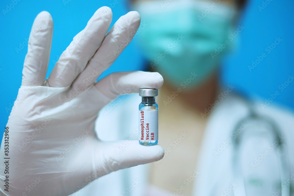 A female Asian physician with surgical mask and white rubber gloves at a clinic, holding a glass bottle of 1 dose HiB vaccine with white background and red letters. A female Asian physician with surgical mask and white rubber gloves at a clinic, holding a glass bottle of 1 dose HiB vaccine with white background and red letters.