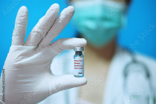 A female Asian physician with surgical mask and white rubber gloves at a clinic, holding a glass bottle of 1 dose MMR vaccine with white background and red letters.