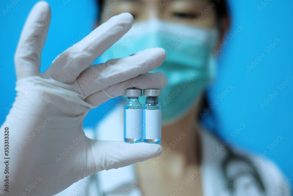 A female Asian physician with surgical mask and white rubber gloves at a clinic, holding two identical glass bottles of vaccine with blank white label. A female Asian physician with surgical mask and white rubber gloves at a clinic, holding two identical glass bottles of vaccine with blank white label.