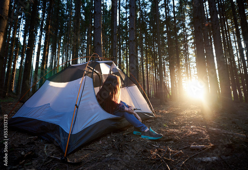 Morning camping in the forest. Young woman hiker sitting in white tourist tent  enjoying sunrise. On background trees and rising sun. Tourism adventure active lifestyle concept
