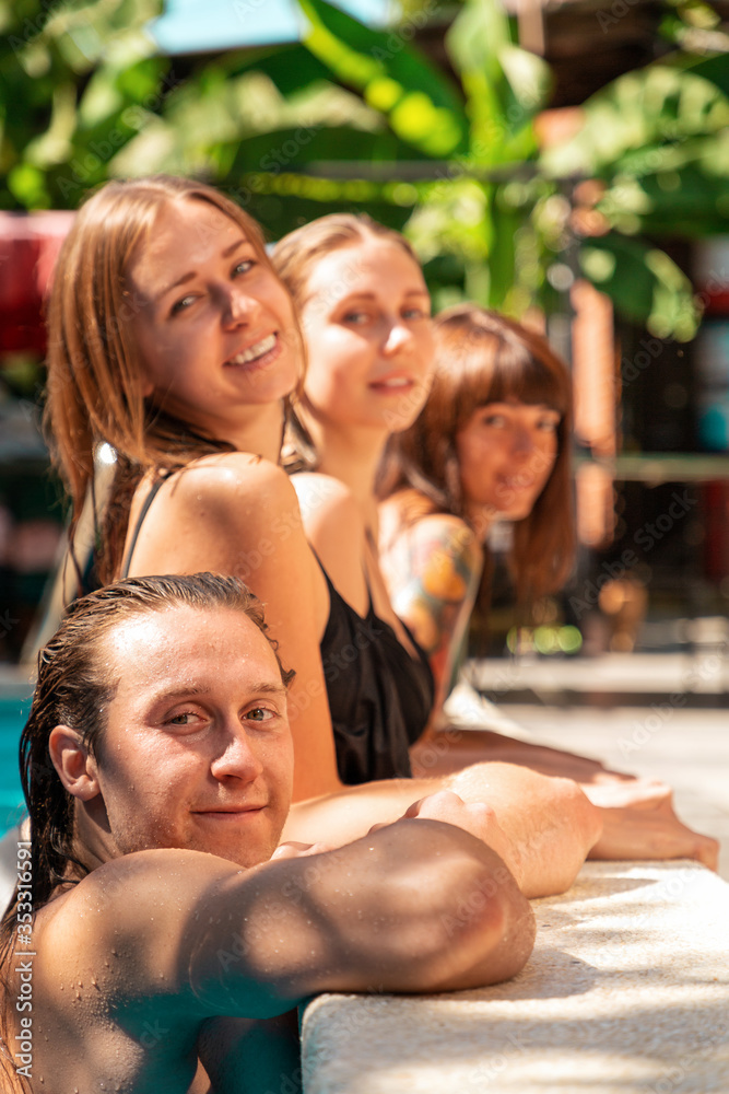 A group of three young women and one man pose in a row, leaning on the edge of the pool.Vegetation is in the background. Summer vacation and friendship concept A group of three young women and one man pose in a row, leaning on the edge of the pool.Vegetation is in the background. Summer vacation and friendship concept