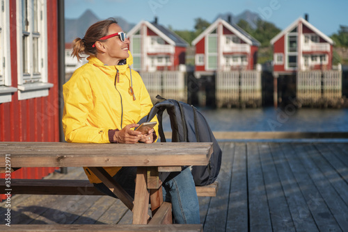 Young woman in a yellow jacket on a background of traditional red Norwegian rorbu houses with a smartphone in hand, traveling to Norway Lofoten Islands © olezzo