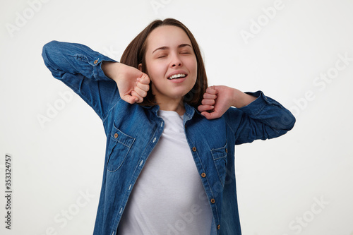 Studio shot of sleepy young good looking brunette woman raising hands while stretching herself and keeping her eyes closed, posing over white background