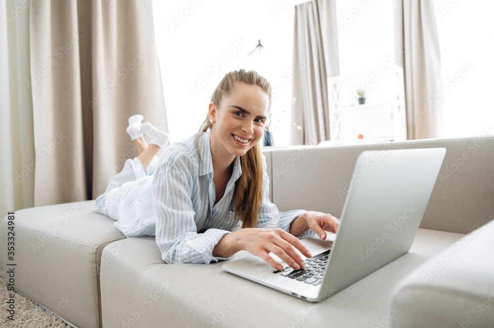 Weekend morning. Young beautiful girl lazily spends time at home on the couch and chatting online with friends in a laptop Weekend morning. Young beautiful girl lazily spends time at home on the couch and chatting online with friends in a laptop