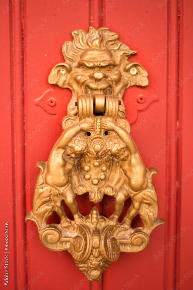 Vertical picture with old style decorated door knob in shape of satire head and two cupidons on red wooden door in Luberon, heart of Provence, France. Medieval ornamental iron antique decoration.