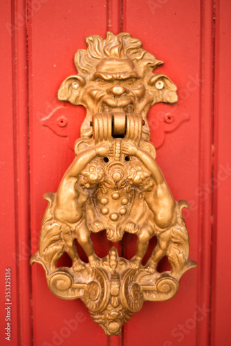 Vertical picture with old style decorated door knob in shape of satire head and two cupidons on red wooden door in Luberon, heart of Provence, France. Medieval ornamental iron antique decoration.