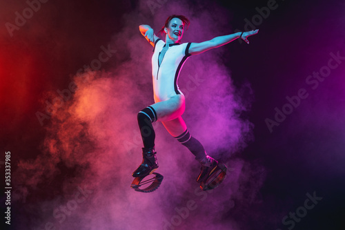 Beautiful redhead woman in sportswear jumping in a kangoo jumps shoes isolated on dark gradient studio background in neon lighted smoke. Active movement  action  fitness and wellness. Slender model.