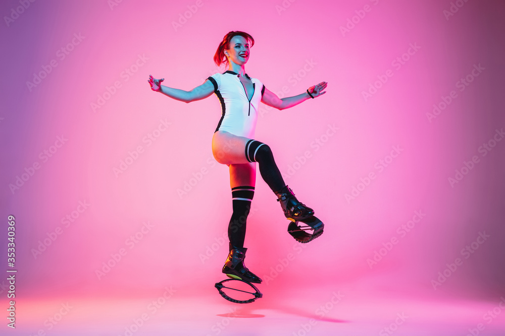 Beautiful redhead woman in sportswear jumping in a kangoo jumps shoes isolated on purple-pink gradient studio background in neon light. Active movement, action, fitness and wellness. Fit female model.