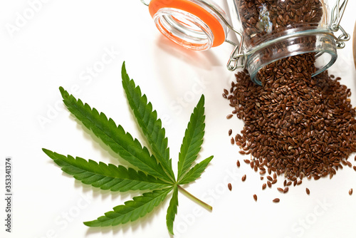Cannabis leaf next to flax seeds spilled out of a glass jar on a white background