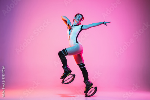 Beautiful redhead woman in sportswear jumping in a kangoo jumps shoes isolated on purple-pink gradient studio background in neon light. Active movement  action  fitness and wellness. Fit female model.