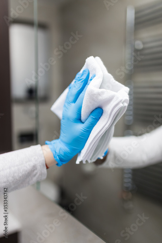 Woman wearing surgical glove and cleaning mirror in bathroom with microfiber cloth.