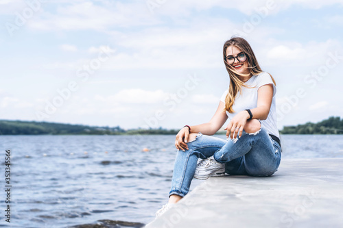 Young woman with long hair in stylish glasses posing on the concrete shore near the lake. Girl dressed in jeans and t-shirt smiling and looking at the camera