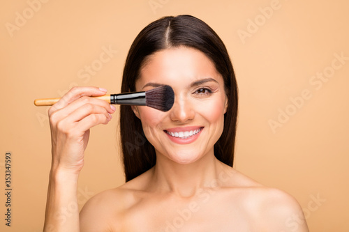 Closeup photo of mature latin naked lady model holding rouge brush hiding one eye applying foundation tone night makeup isolated beige pastel color background