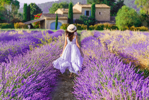 Provence, France. Charming young woman in Blooming Lavender fields at background of beautiful traditional French Provencal house.  Back view of lovely lady wearing waving Boho Chic purple Dress, hat. photo