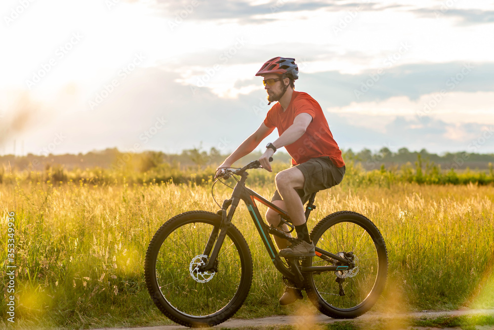 A cyclist in a bright orange T-shirt rides a bicycle along the meadow during sunset. A cyclist in a bright orange T-shirt rides a bicycle along the meadow during sunset.