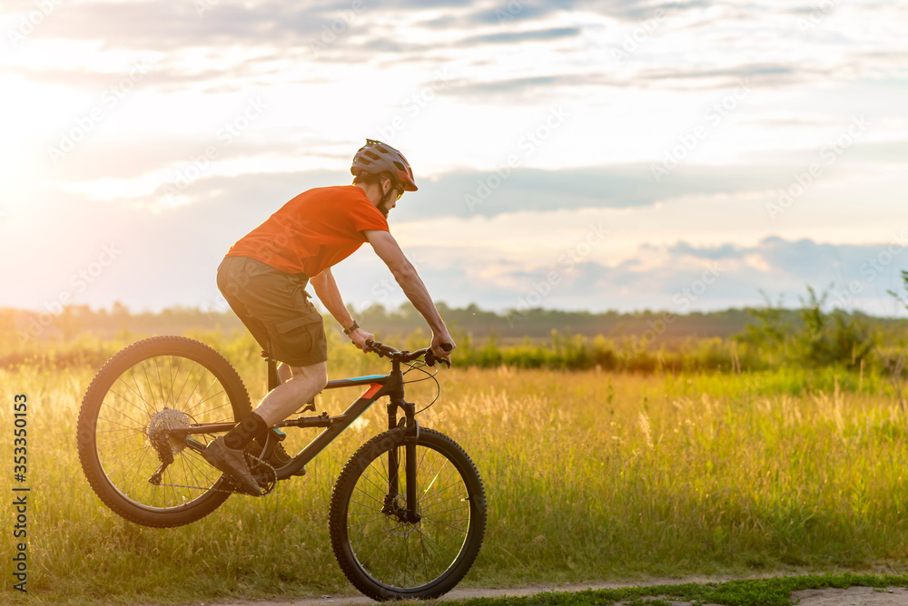 A cyclist in a bright orange T-shirt stands on the front wheel of a bicycle along the meadow during sunset. A cyclist in a bright orange T-shirt stands on the front wheel of a bicycle along the meadow during sunset.