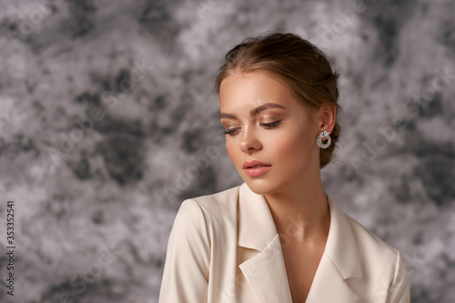 Closeup beauty face portrait of young elegant woman with perfect makeup and hairstyle in white blazer against gray textile background photo