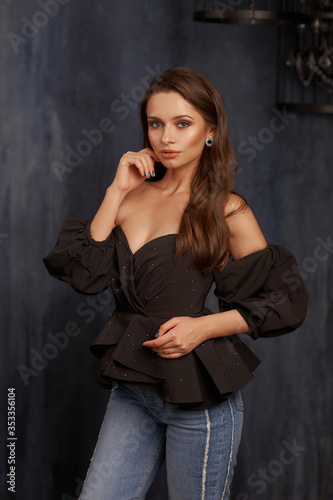 Studio portrait of young elegant woman with hairstyle and makeup in dark interior with blue walls. Pretty girl standing and posing