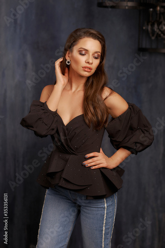 Studio portrait of young elegant woman with hairstyle and makeup in dark interior with blue walls. Pretty girl standing and posing