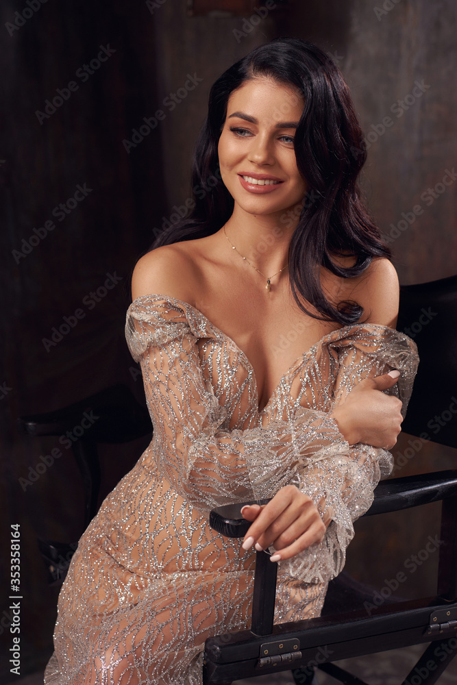 Young beautiful elegant pregnant woman wearing luxury expensive evening dress, sitting at high chair and posing alone in dark stylish interior
