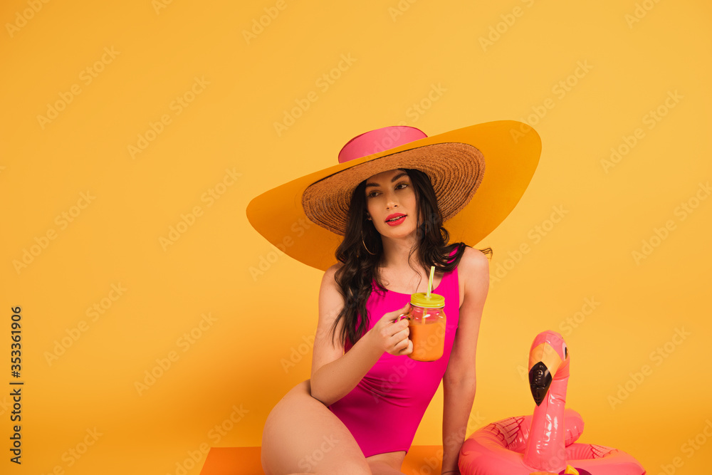 beautiful girl in straw hat and swimsuit holding glass with orange juice near inflatable ring on yellow beautiful girl in straw hat and swimsuit holding glass with orange juice near inflatable ring on yellow