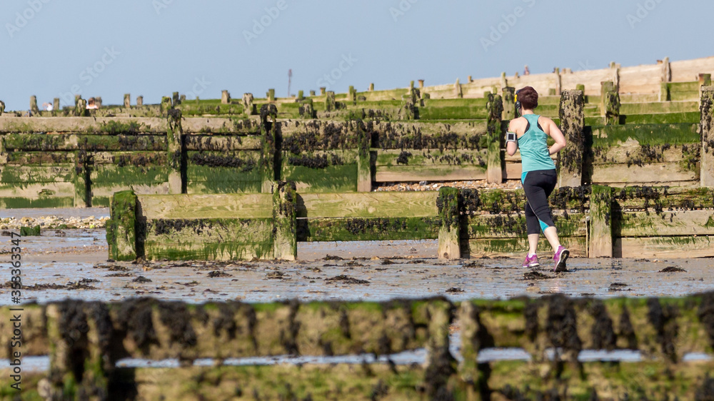 Rear View of Adult Woman Running Along a Beach Wearing Headphones.  Several Wooden Groynes are Visible.  There is a Blue Sky