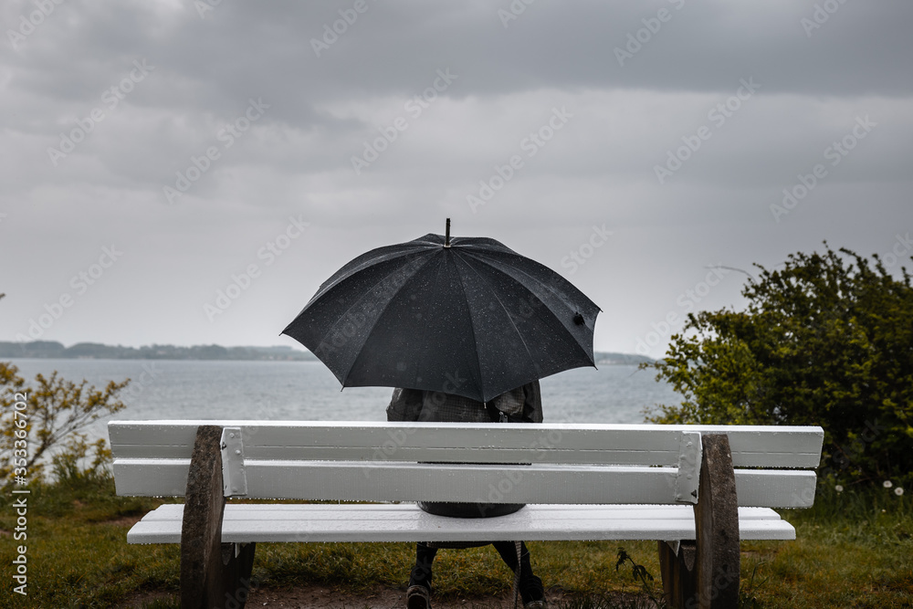 Young Women in rainjacket with umbrella sitting on bench at coastline in rainy weather
