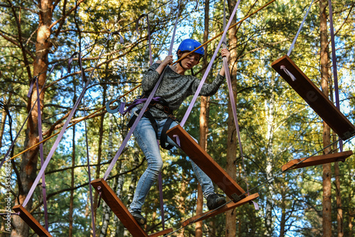 In a rope park, a beautiful young girl overcomes a difficult obstacle at high altitude, a shaky structure, a little scary and exciting, against the backdrop of huge trees and a clear sky, in a pine fo