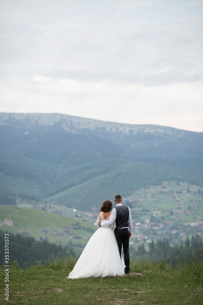 Wedding. The bride and groom running through the mountain slope. After wedding session. The guy in the white shirt and waistcoat, and a girl in white dress walk on the green glade in the mountains