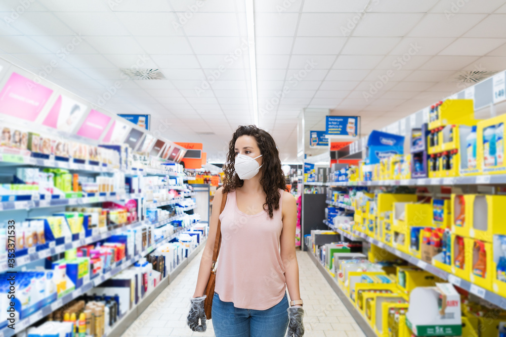 Caucasian woman wearing face mask and rubber glove push shopping cart in supermarket department store. Girl choosing, looking grocery things to buy at shelf during coronavirus crisis outbreak.