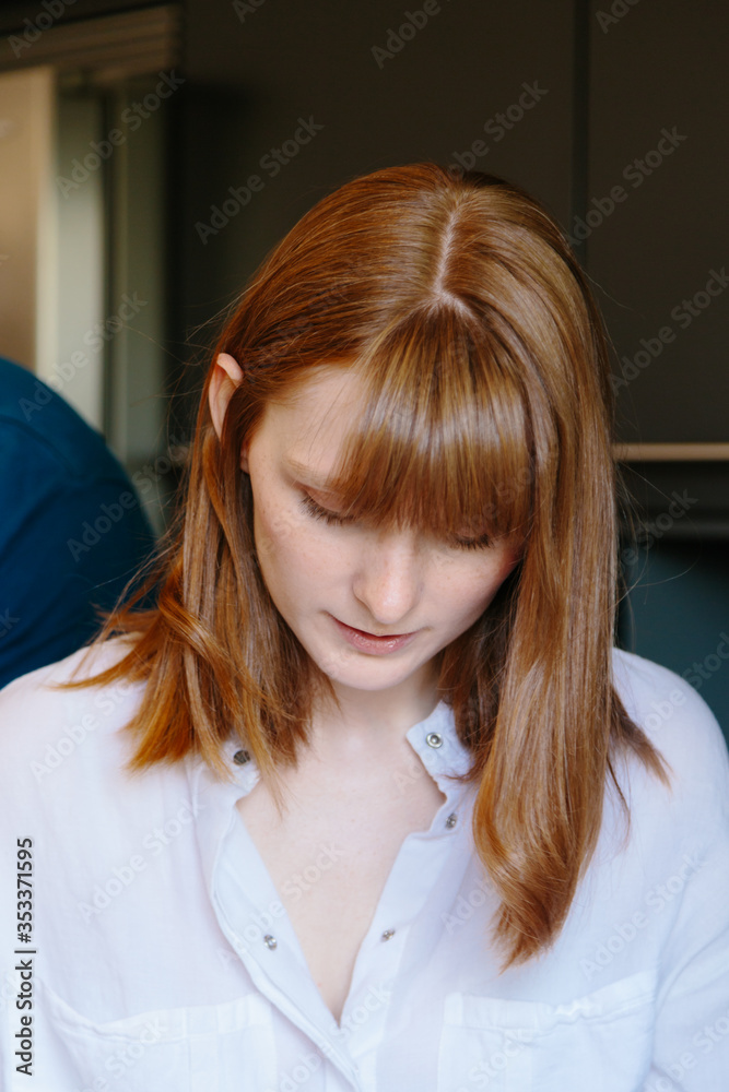 A vertical portrait of a young redheaded woman with a white shirt facing the floor