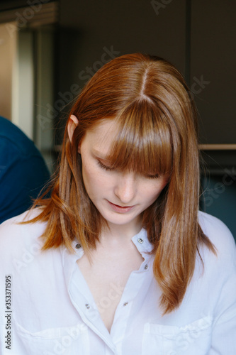 A vertical portrait of a young redheaded woman with a white shirt facing the floor