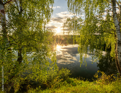 Scenic view at beautiful spring sunset on a shiny lake with green branches  birch trees  bushes  grass  golden sun rays  calm water  deep blue cloudy sky and forest on a background  spring landscape
