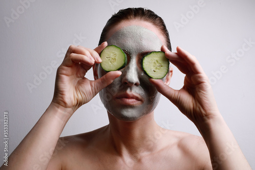 Portrait of young woman with a face mask on holding a cucumber slices in front of one eye. Beauty and skincare concept.