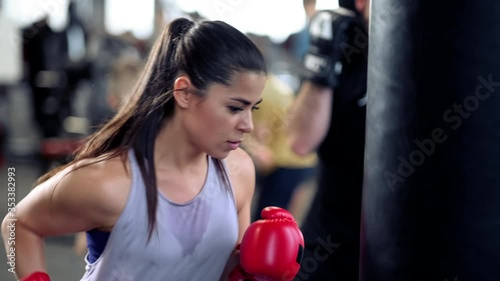 A beautiful athletic young brunette woman in sportswear and red boxing gloves trains bumps on a punching bag in a fitness gym.