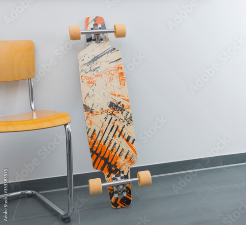 A drop through longboard skateboard is leaning against a white wall next to a wooden cantilever chair in a modern apartment with a granite floor. Location shot with copy space, no people.  