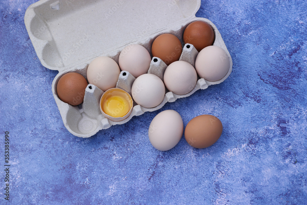 Chicken eggs in a carton on a blue background. One egg is broken. Horizontal banner with place for text. View from above
