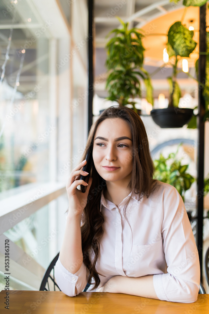 Young happy woman talking on mobile phone with friend while sitting in modern coffee shop