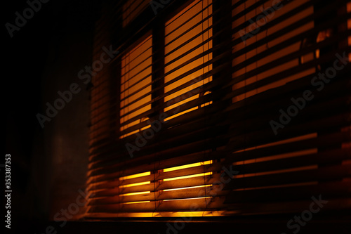 Dark, moody atmosphere set by yellow light softly piercing through the blinds. The shutters create a contrast between light and shadow. Concept of mafia, fear, kidnapping, sleep, covid19, confinement.