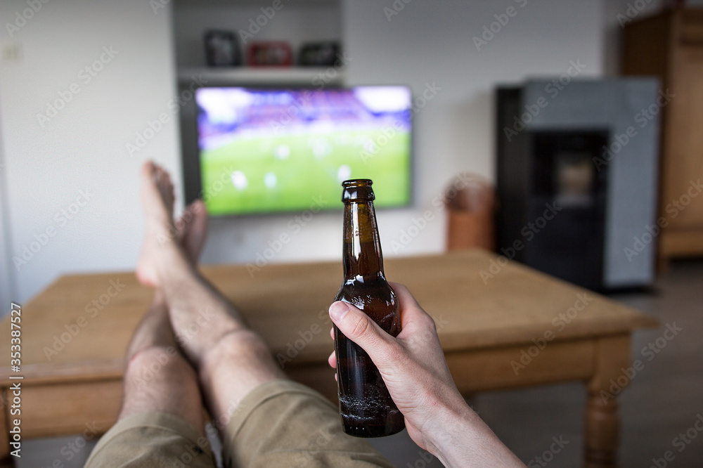 A person's hand holding a brown beer bottle, whil watching a soccer match  on tv. His legs are placed on the wooden table to relax. Stock Photo