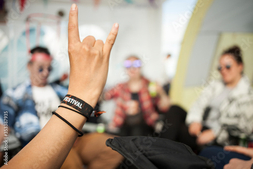 A hand showing the sign of the horns, whichs usually refers to the appreciation of rock music. The wristband says 'Festival'. A group of friends is sitting, chatting and drinking in the background.