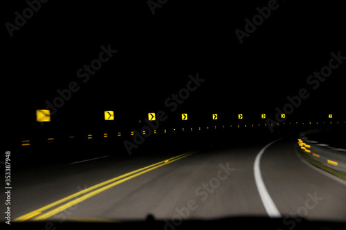 A series of yellow reflective 'sharp turn' signs being lit up by the lights of an oncoming car. The pitch black night sky is in contrast with the signs and street. Concept of speed and car accidents.
