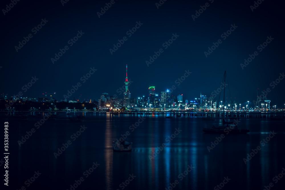 The lights of Auckland city downtown and the port reflected in Okahu bay with a small boat in the foreground. Soft focus