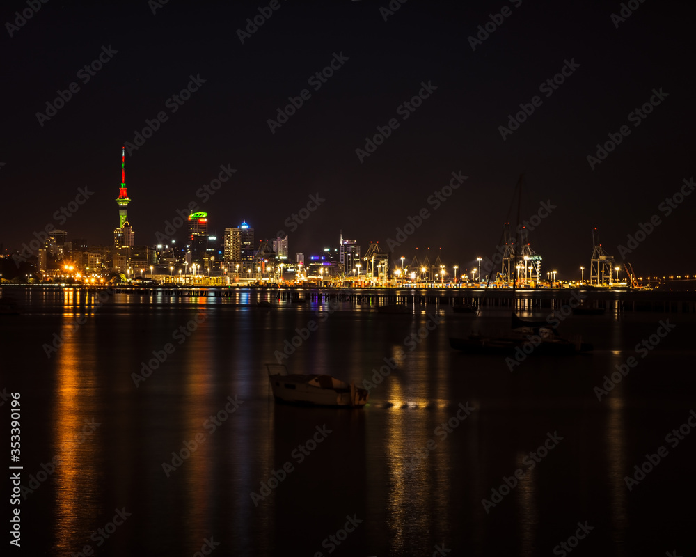 Bright lights of Auckland city downtown and the port reflected in Okahu bay with a small boat in the foreground. Soft focus