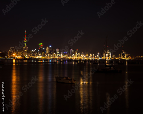 Bright lights of Auckland city downtown and the port reflected in Okahu bay with a small boat in the foreground. Soft focus