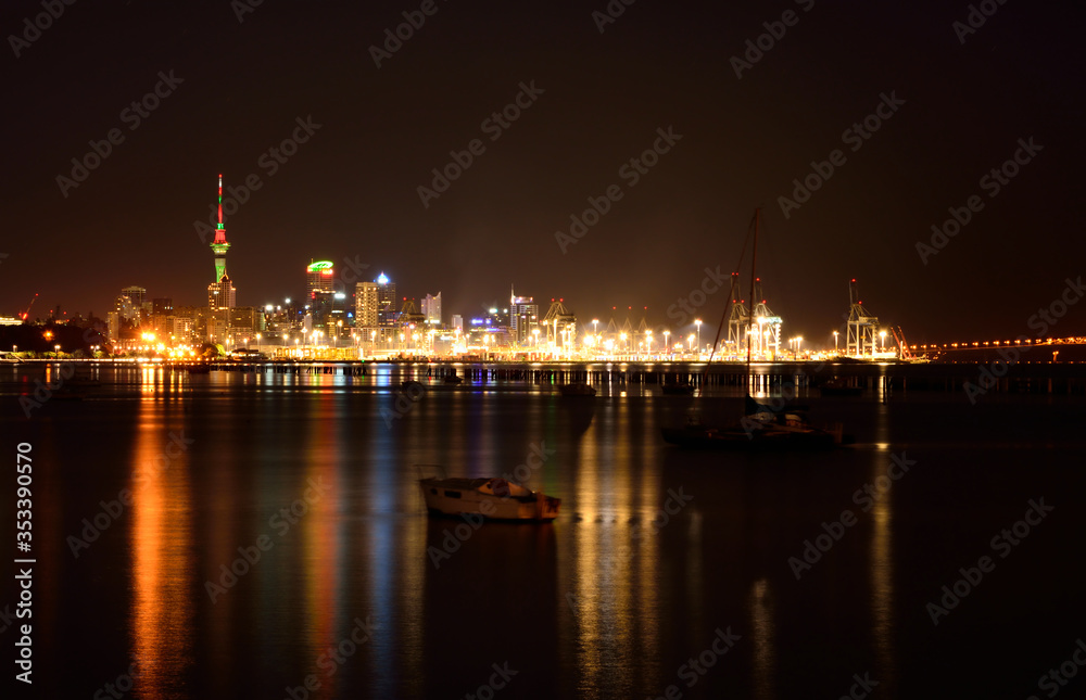 Midnight in Auckland. The lights of city downtown and the port reflected in Okahu bay with a small boat in the foreground. Soft focus